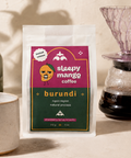Sleepy Mango Coffee Burundi package on a beige surface with a plant and coffee maker in the background
